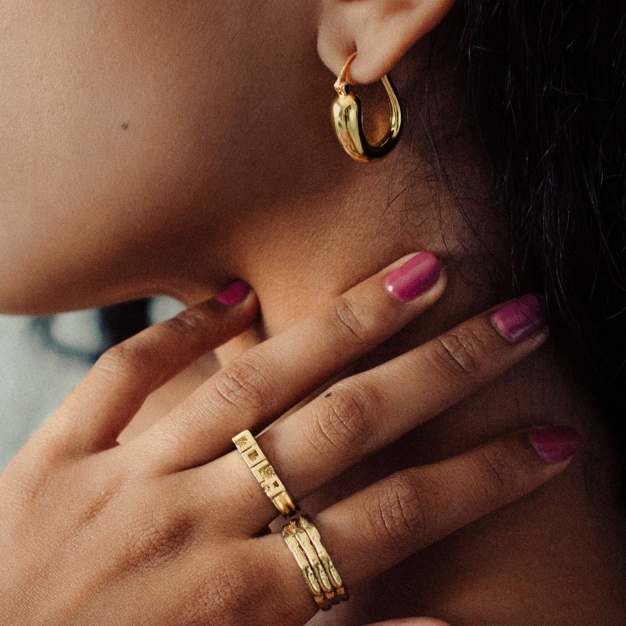 Close-up of a person wearing gold hoop earrings, rings, and pink nail polish. iris 1956 jewelry